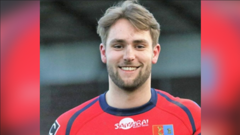 Jon Devereux is seen from his shoulders up wearing a red rugby uniform. He is smiling.
