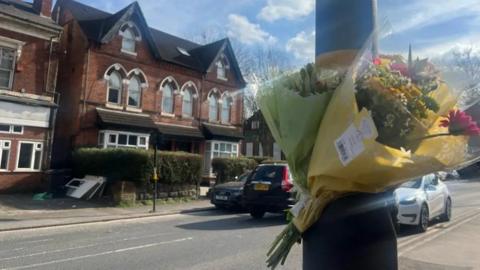 Two bouquets of flowers attached to a grey lamppost on a residential street. There are a few cars in the background.