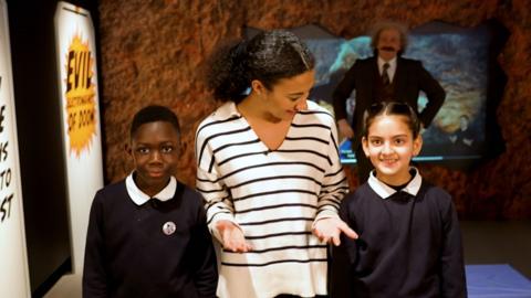 Presenter Nina with two school children in an exhibition, with an actor playing Albert Einstein in the background