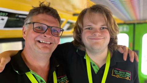 Jason and Luke stoner standing inside the bus which has a yellow and green paint scheme. Jason has glasses and luke has long brown hair. Both are wearing black t shirts with the logos Retro Reset