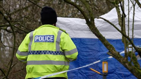 A police officer standing outdoors in what appears to be a wooded or rural area. The officer is photographed from behind and is wearing a bright yellow high‑visibility jacket with the word “POLICE” clearly displayed on the back. They are also wearing a black beanie hat. In front of the officer, a large blue‑and‑white tent is set up. Striped police tape is visible in the foreground, marking off the area."
