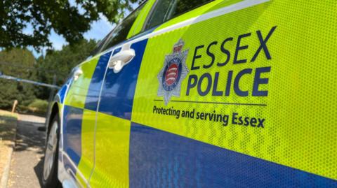 A police car is parked outside a fence. The fluorescent yellow and blue markings are topped with a logo which says Essex Police, Protecting and serving Essex.