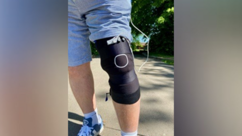 A black sleeve strapped on to a mans knee, with a white wire leading to a pocket in the man's blue shorts