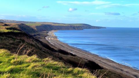 The beach at Horden backed by dark bluffs topped with green grass. The land and beach curves out into the horizon next to a very blue sea under light blue skies.