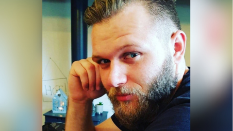 A picture of Luke Davis inside a house. He is leaning on a counter with his right arm and looking at the camera. There are some home decorations such as a plant in a pot and a mini house on a desk.