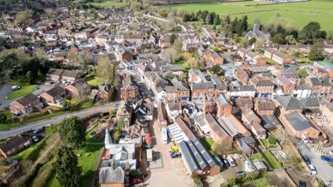 An aerial view of Tenbury Wells in the Malvern area. The small town is surrounded by green fields and the buildings are largely red brick and between two and three storeys high.