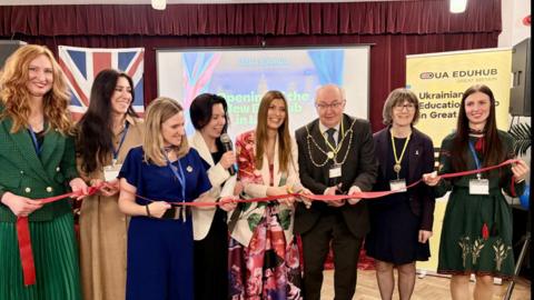 Seven women in formal outfits hold a red ribbon while a man in a black suit and mayoral chains cuts the ribbon.