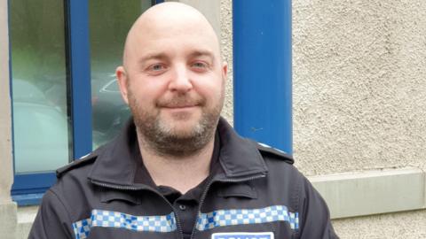 A head and shoulder picture of Inspector Peter Aiston. He is wearing a black police jacket with blue and white check markings. He is bald with a short trimmed beard. He has blue eyes. He is looking at the camera and slightly smiling. Behind him is the wall of a building with a window with a blue frame.