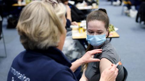 A woman in a blue face mask having her sleeve rolled up by another person, who is holding a needle.