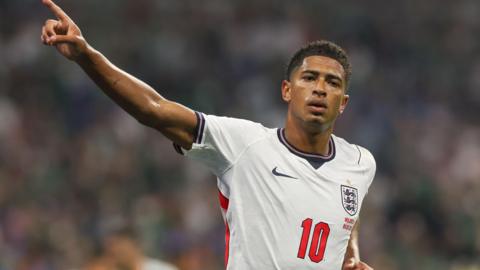 Jude Bellingham celebrates a goal with one arm out, his index finger pointed to the sky. He wears a white England shirt with a red number 10 on it.