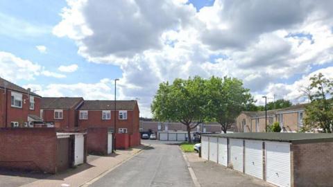 A small street with houses and garages to the left, and a block of garages with white doors to the right