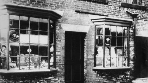 Black and white image of two lattice window fronts showcasing wares on either side of an entrance door.