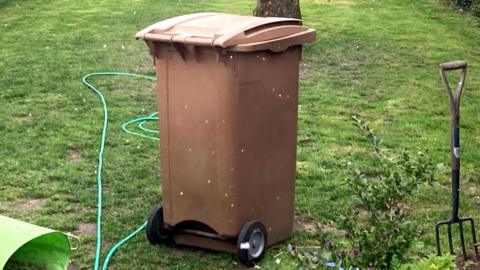 A brown garden waste bin sits in a garden. A green hose pipe lies on the grass around it as well as a garden fork.