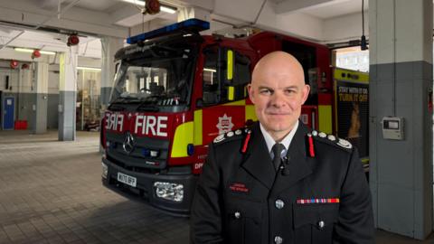 The London Fire Commissioner - a bald male in ceremonial uniform, a black suit jacket with badges on the shoulders. A parked red firetruck is present in the background of the shot.