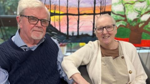 Sally and Kevin are looking at each other and smiling. Kevin is wearing a blue and white striped shirt and a navy knitted tank top. Sally is wearing a cream cardigan, brown t-shirt and light brown dungarees. Both have short white hair and glasses.