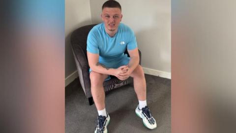Dale Martin is pictured sitting on a chair wearing a light blue t-shirt, shorts and white trainers.