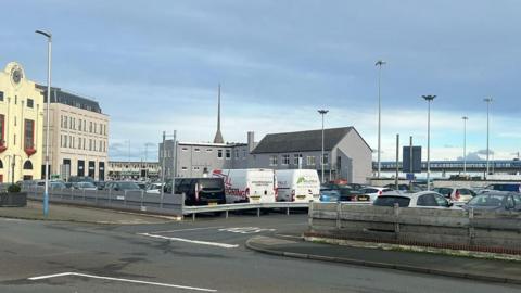 The site of the proposed development, it is an existing car park. Cars and vans, a range of colours such as blue, black and silver, line up.