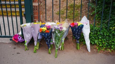 A row of about a dozen bouquets of flowers are resting against school gates. 