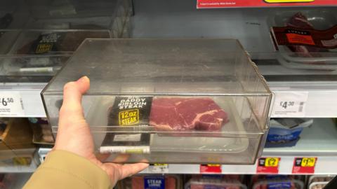 A sirloin steak in normal plastic packaging individually inside a clear durable plastic box with a security tag on the side.