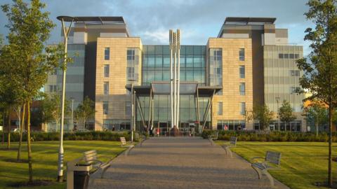 The computer-generated image of the exterior of Peterborough City Hospital. A large glass-panelled building with a central walkway with green space on either side. The walkway has benches and is lined with trees. 