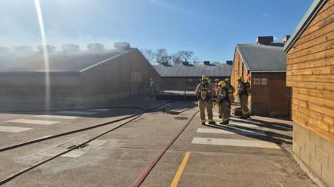 A number of firefighters stand on a courtyard in the middle of a farm site with several buildings around them. The building on the left is emanating white smoke.