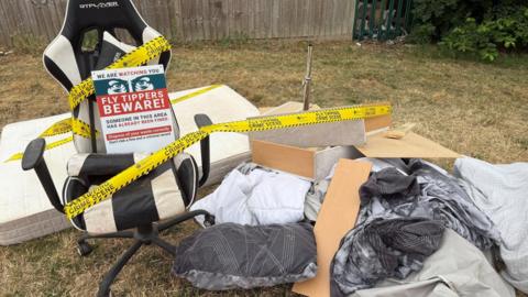 A large pile of rubbish, including bags of waste, and a large chair, with yellow "fly tipping - crime scene" tape all around it and a large sign. It is all sat on grass. A fence is behind the rubbish.