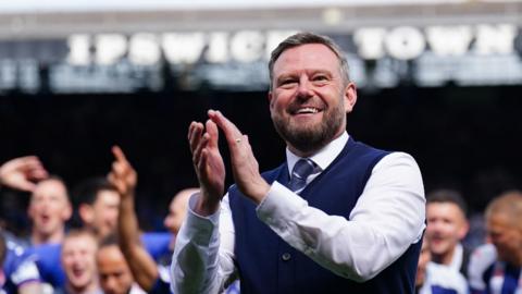 Mark Ashton wearing a white shirt, with a blue tie and waistcoat. He is clapping his hands and is inside a football stadium. The words "IPSWICH TOWN" are behind him on the top of a grandstand.
