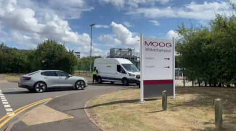 A road leading to a fenced off area with a silver car and a white van on it and a large white sign with the words 'Moog Wolverhampton' on it