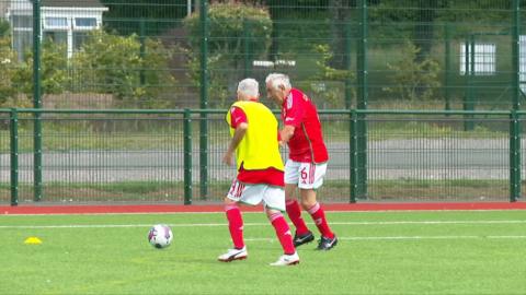 Two veteran footballers, both in red shirts, one with a yellow bib