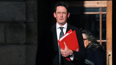 Enoch Burke coming out of a building. He's holding a red file. He wears galsses and has a black suit on with a white shirt and a stripy tie.
