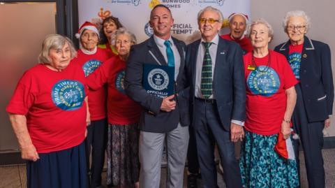 A group of elderly choir members gathered around Tony Christie and Carl Saville, who is holding a Guinness World Record certificate. 