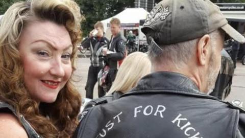 Hev Cagnasso with long brown curly hair smiling at the camera in close-up next to a man wearing a dark-coloured cap and a leather jacket with the words "JUST FOR KICKS" in white writing. Two bikers are visible in the background - one is drinking from a shite cup. There is a marquee behind them.