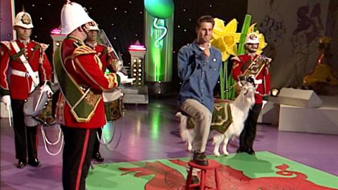 Presenter Tim Vincent preparing to eat a raw leek in front of Welsh soldiers in formal dress.