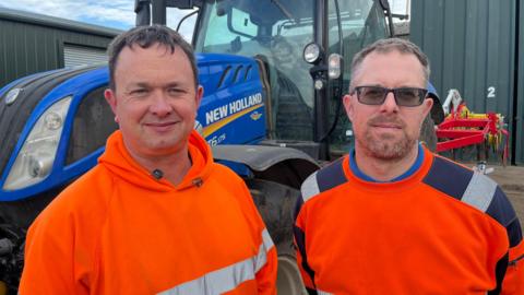 Farmers Mat and Lloyd Smith wear high-vis orange jackets and there is a tractor behind them.
