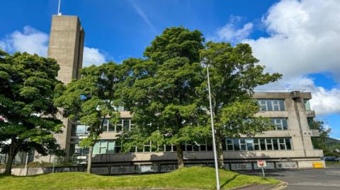 A grey concrete council headquarters building which is concealed by trees on a bright sunny day with just a few clouds with a blue sky