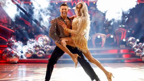 A man and woman dancing while on the Strictly Come Dancing stage. There are red lit steps behind them with giant silver baubles down either side. The man is wearing a sparkly shirt and black trousers and he is holding the woman's left leg across his body. The woman has long light blonde hair and is wearing a short golden dress with tassles. 