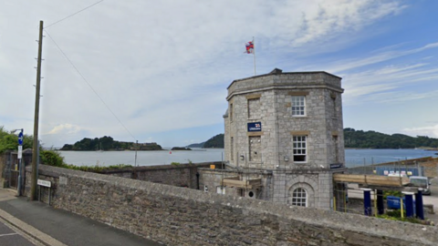 Exterior of a lifeboat station. A small brick wall runs alongside a pavement. There is a stone grey building with multiple windows and lifeboat signage displayed on its outside wall. A calm sea is in the background. Trees are in the distance.
