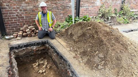 A man wearing a high vis jacket and hard hat is kneeling next to a small pit with a large pile of dirt nearby