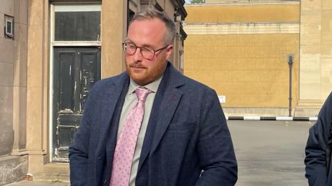 A man with short, light brown hair and a short ginger beard leaves court. He is wearing a pink tie, light shirt and grey jacket. 