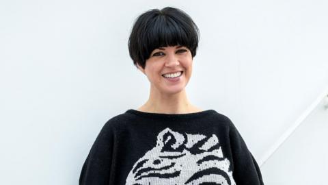 Dawn O'Porter stood in front of a white background. She has a black and white jumper on and a short black bob hairstyle.