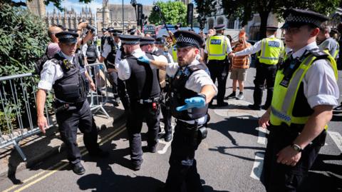 A number of police officers on a road with some members of the public taking photos and filming. The police appear to be carrying a person although only their foot can be seen in the image.