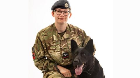 A woman wearing an RAF Police uniform, including a black beret and camouflage jacket, with a black dog by her side.