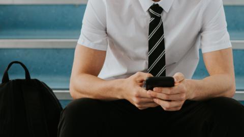 A stock image of a boy sitting on blue stairs. He is wearing a white short-sleeved shirt, with a black and white tie and black trousers. He is holding a phone in both hands. A black rucksack is sitting on the stairs on the left of the image.