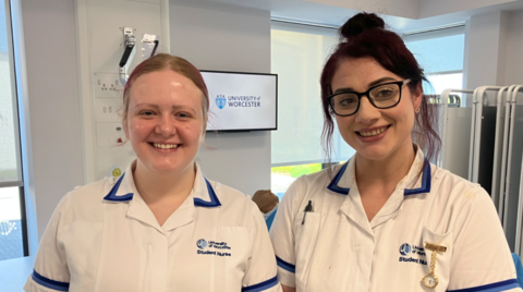 Two women in a mainly white uniform including the words University of Worcester Student Nurse. Windows and walls are behind them and there is a screen on the wall featuring the words University of Worcester.