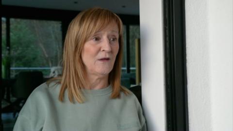 Dianne Morgan, a woman with short red hair in a green jumper, stands near a window in her house