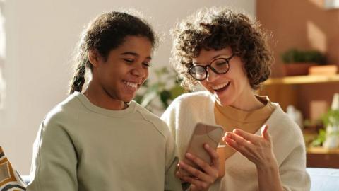A girl and a woman smile while looking at a smartphone together