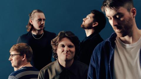 A promotional image of the band which is made up of five young men with dark hair. The man at the front on the right is looking down and is wearing a white t-shirt and blue shirt over the top. The man in the middle has longer brown hair and is scrunching up his face. On the left is a man with his back to the camera while he looks off to the to the left. He is wearing a blue striped t-shirt. Behind his is a man with brown long hair which is half tied up. He is wearing a black t-shirt and a chain necklace. To the right is a man with dark hair and a beard looking at the ceiling.