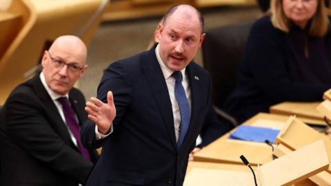 Neil Gray, a bald man in a dark blue suit and dark blue tie with a white shirt, speaks while looking to his right in the Scottish Parliament chamber 