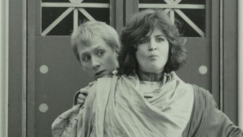 Actor Ruth Jones is pictured standing in front of another student in a performance of Casina by Plautus in 1988. She has dark hair and is wearing robes with a male actor hiding behind