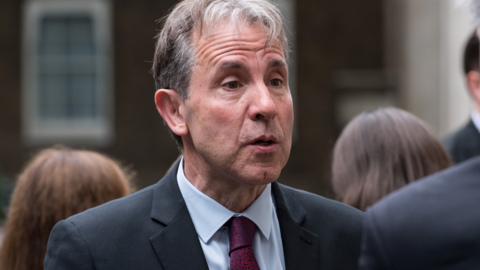 Dan Norris speaking to someone while wearing a navy suit and red and blue tie. He is stood outside with people behind him.